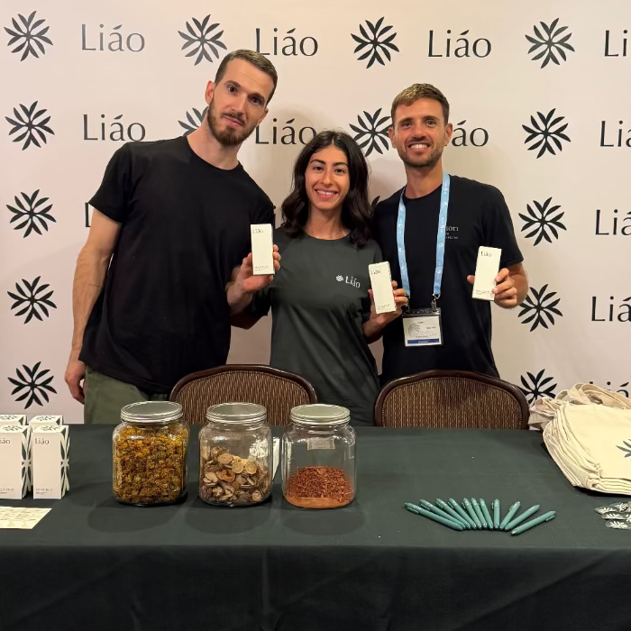 Three people standing behind a table with jars and promotional materials, against a backdrop with 'Liáo' branding.